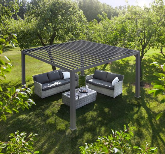 De Lamellen overkapping Handbediend met een lamellendak staat in een groene tuin en biedt schaduw aan twee grijze buitensofa's en een bijpassende salontafel, omringd door weelderige bomen en zonlicht.