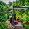 Vier personen zitten en één staat onder de lamellenoverkapping Handbediend in een weelderige tuin, gezellig rond een rode tafel. Groen en tuinmeubilair maken de uitnodigende, ontspannen sfeer compleet.