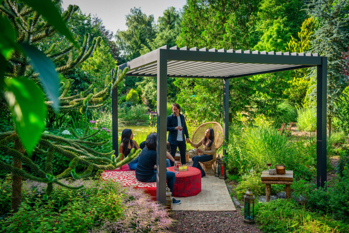 Vier personen zitten en één staat onder de lamellenoverkapping Handbediend in een weelderige tuin, gezellig rond een rode tafel. Groen en tuinmeubilair maken de uitnodigende, ontspannen sfeer compleet.