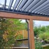 De Lamellen overkapping Handbediend, een houten pergola met verstelbare lamellen, staat op een bewolkte dag bij een kleine tuinvijver, omgeven door groene planten en een houten omheining.