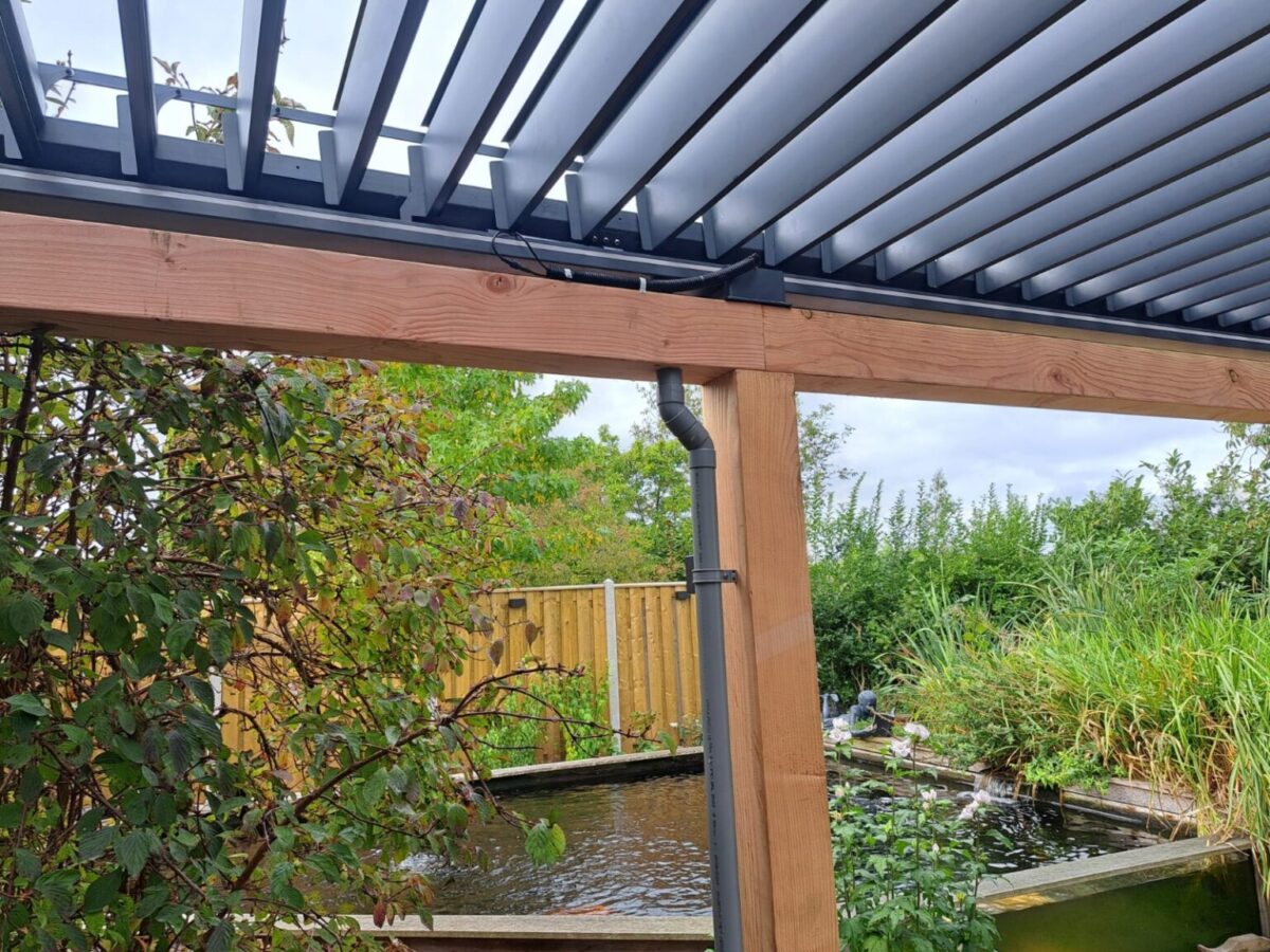 De Lamellen overkapping Handbediend, een houten pergola met verstelbare lamellen, staat op een bewolkte dag bij een kleine tuinvijver, omgeven door groene planten en een houten omheining.