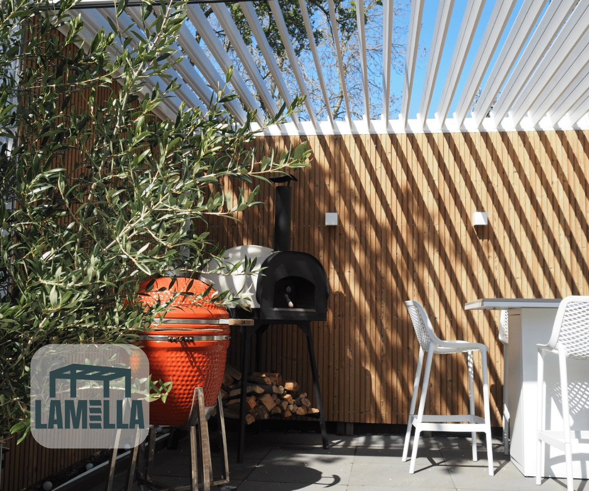 Houten wanden omringen een buitenpatio met gestreepte schaduwen van een witte pergola. Een rode keramische grill en een zwarte pizzaoven staan links, met daaronder opgestapelde houtblokken. Rechts staan witte barkrukken en een tafel. De Lamellen overkapping Elektrisch biedt verstelbare schaduw, terwijl er een kleine olijfboom bij de grill staat.