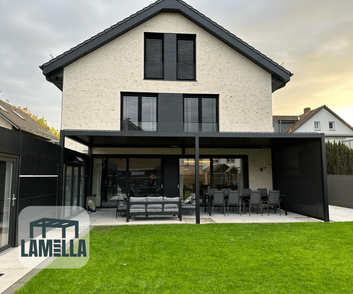 Een modern huis met twee verdiepingen, een lichte bakstenen buitenkant en zwarte accenten, heeft een groot terras met zitplaatsen en een eettafel onder de Lamellen overkapping Elektrisch. De tuin is weelderig met groen gras en het LAMELLA-logo staat in de linkerbenedenhoek.