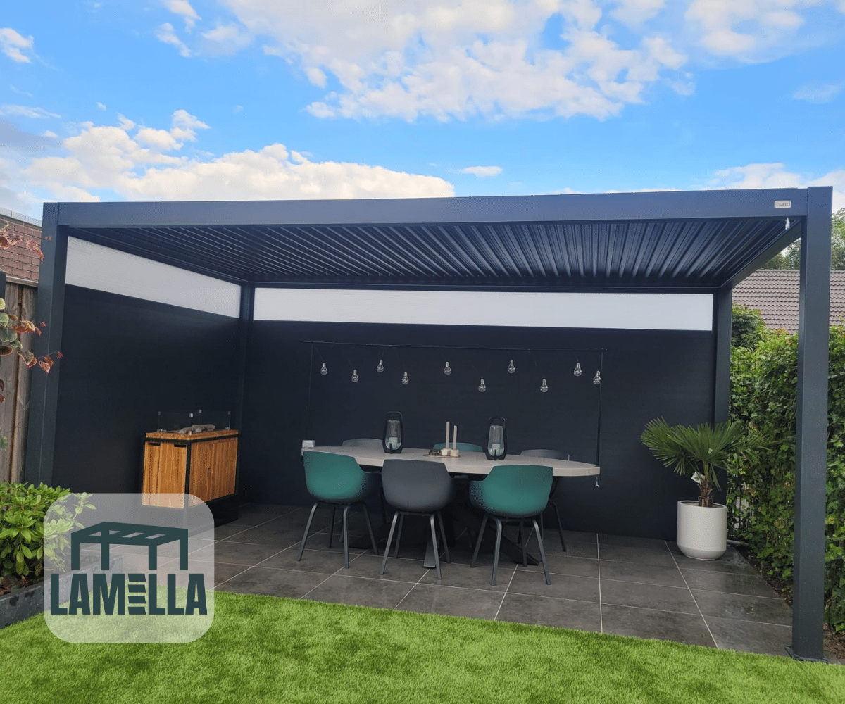 Buitenpatio met een moderne pergola met een zwart frame. Daaronder beschut een elektrische Lamellen overkapping Elektrisch een eethoek met tafel, stoelen en hanglampen. Links staat een houten opbergkast. De lucht is gedeeltelijk bewolkt. LAMELLA-logo in de hoek.