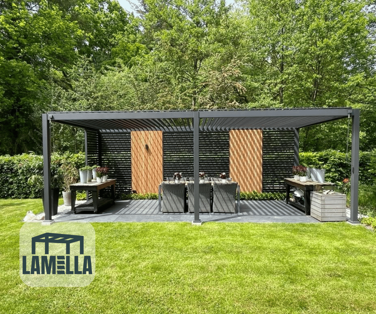 Een moderne Lamellen overkapping Elektrisch met een lamellendak staat in de weelderige tuin, met twee tafels en stoelen tussen levendige potplanten. Het groene gras en de bomen vormen een verbluffende achtergrond, met het Lamella-logo subtiel in de linkerbenedenhoek.