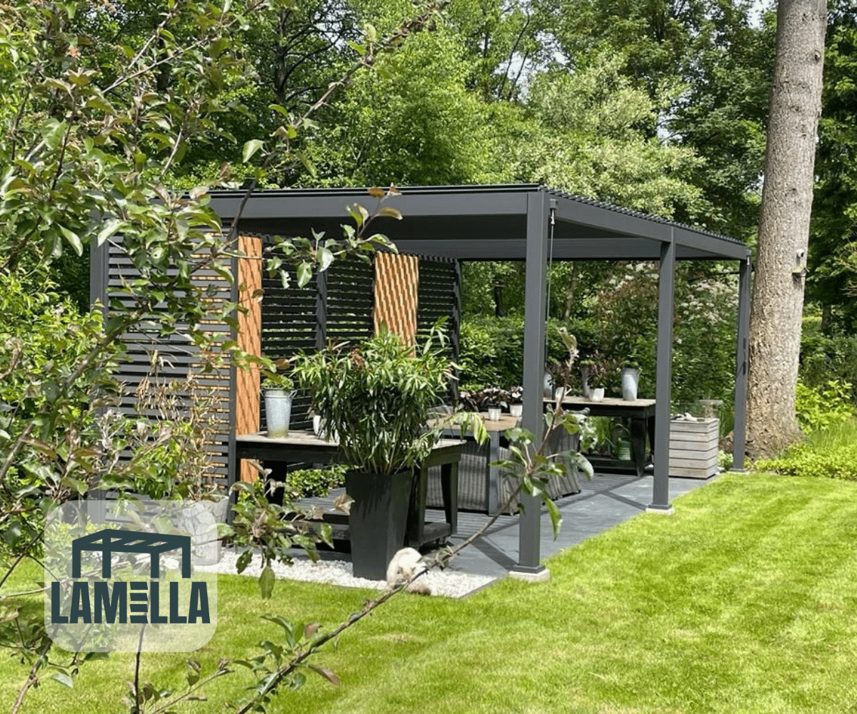 Een moderne pergola met donker metalen frame en houten lamellen, de Lamellen overkapping Elektrisch, siert een grijs terras met een eethoek te midden van weelderig groen. In de buurt rust een kleine witte hond op het gras. Het LAMELLA-logo verschijnt linksonder.