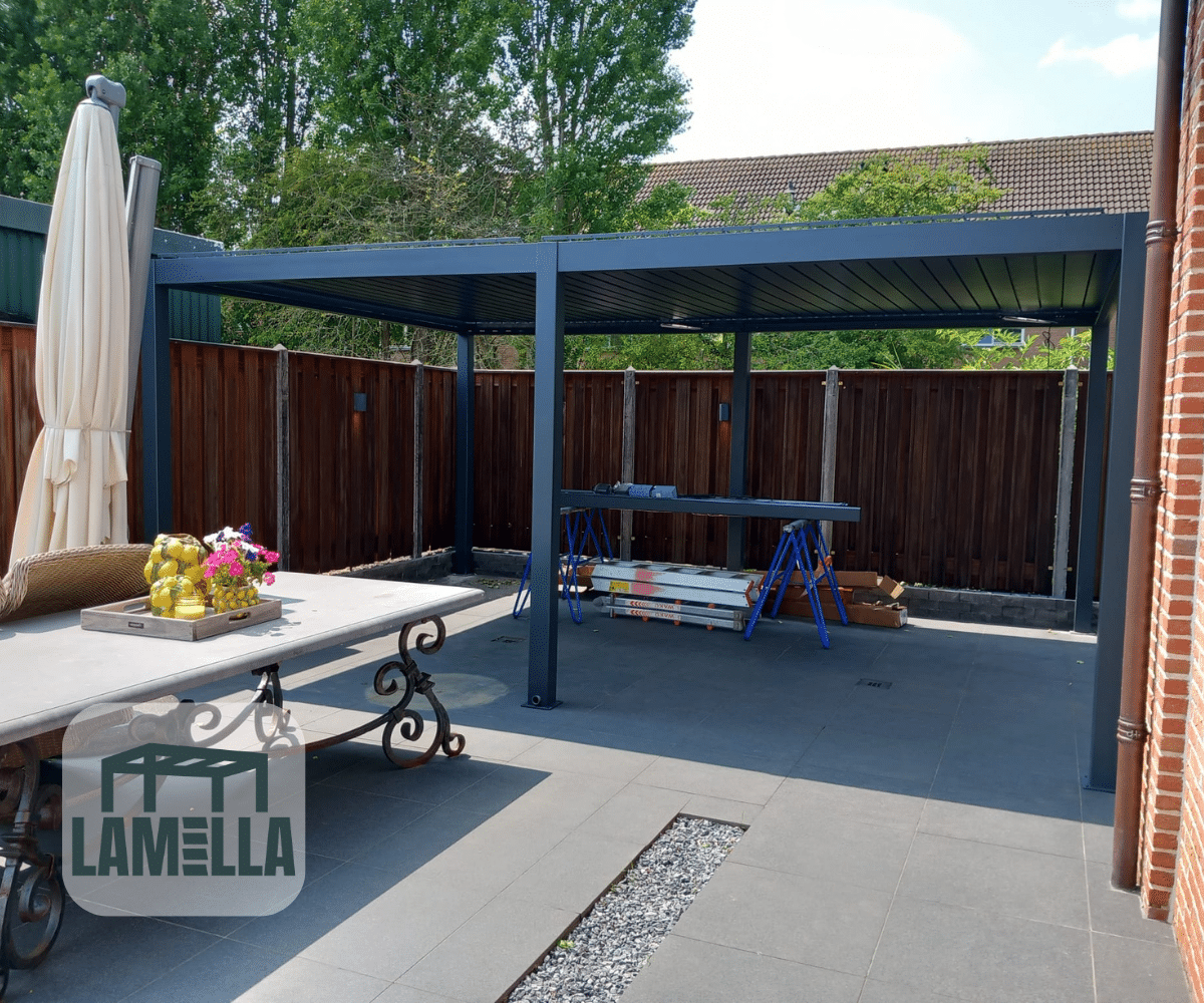 Een buitenpatio toont een pergola met een tafel met fruit en een vaas. Op de achtergrond is een bouwwerkstation op zaagbokken te zien. Hoge bomen en een houten hek omarmen het gebied, terwijl de moderne Lamellen overkapping Elektrisch flair toevoegt, met het LAMELLA-logo prominent op de voorgrond.