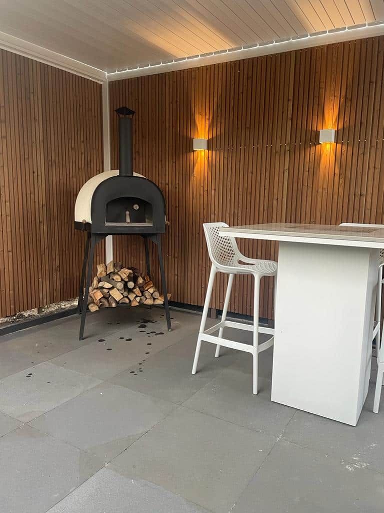 Een klein terras met een houtgestookte pizzaoven aan de linkerkant, daaronder gestapelde houtblokken. Een hoge witte tafel en een bijpassende kinderstoel staan aan de rechterkant. De houten muren hebben twee warme lampen en de vloer is van grijze tegels.