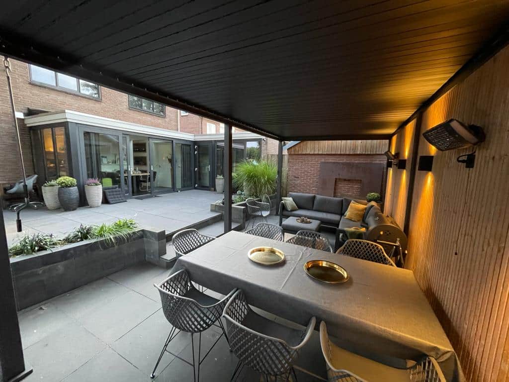 Modern buitenterras met een overdekte zithoek. Beschikt over een eettafel, stoelen en een bankstel. Sfeerverlichting en potplanten zijn aanwezig. Het aangrenzende huis heeft grote ramen en schuifdeuren.