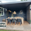 Buitenpatio met een houten eettafel en zes zwarte stoelen onder een pergola. De ruimte is versierd met een hangende rieten lamp, verschillende muurornamenten en een ingelijst portret. Een logo met het woord Lamella staat in de hoek.