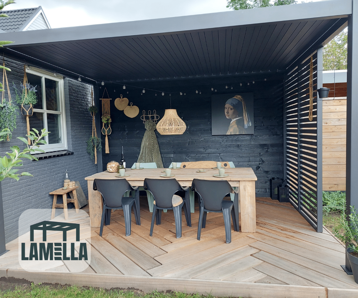 Buitenpatio met een houten eettafel en zes zwarte stoelen onder een pergola. De ruimte is versierd met een hangende rieten lamp, verschillende muurornamenten en een ingelijst portret. Een logo met het woord Lamella staat in de hoek.