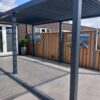 Een modern buitenterras heeft een strakke metalen pergola die schaduw biedt over een betegelde vloer. Een houten hek met strandthemakunst en potplanten langs één kant. Glazen deuren van een bakstenen huis zijn zichtbaar op de achtergrond onder een heldere lucht.