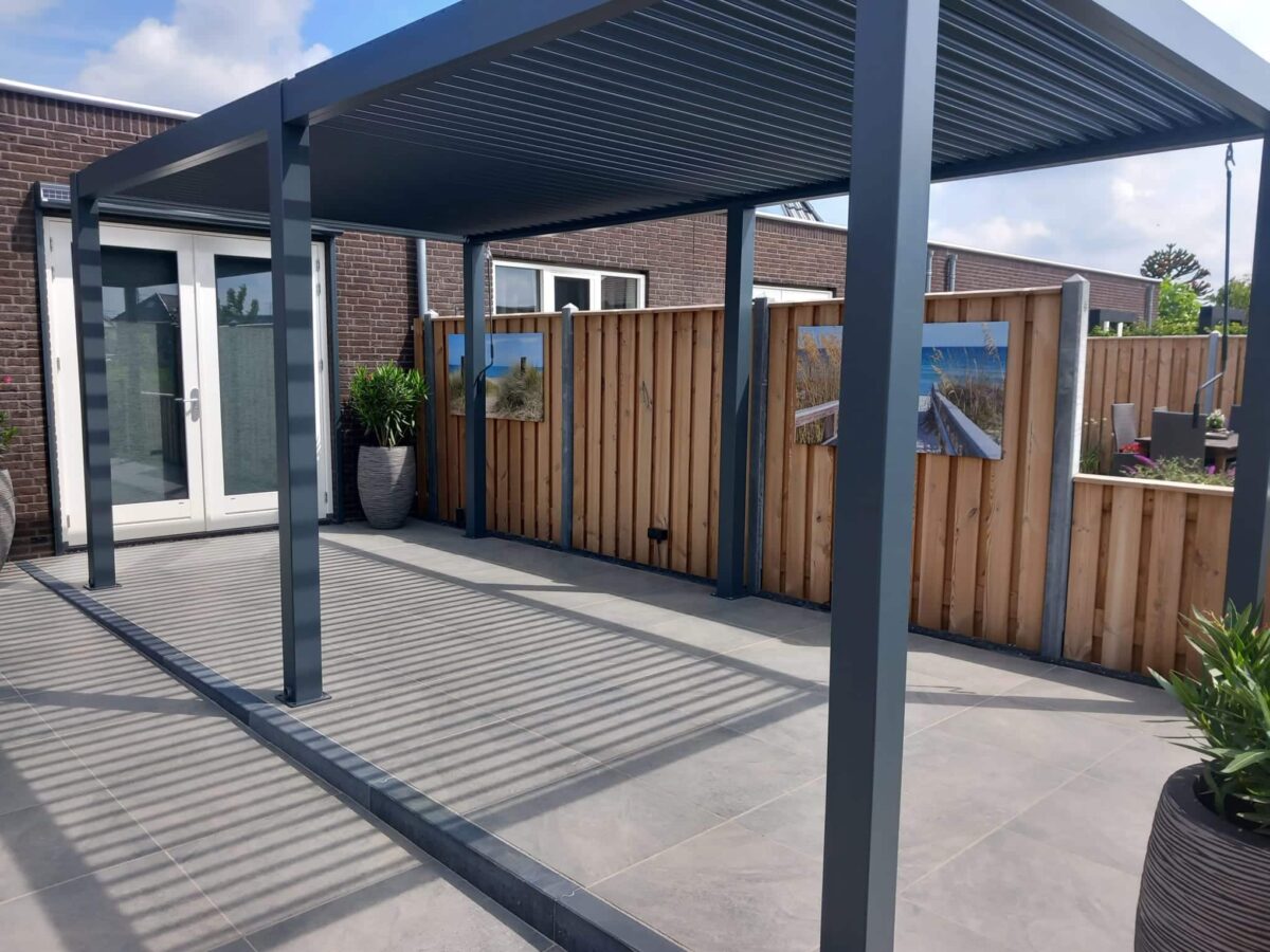 Een modern buitenterras heeft een strakke metalen pergola die schaduw biedt over een betegelde vloer. Een houten hek met strandthemakunst en potplanten langs één kant. Glazen deuren van een bakstenen huis zijn zichtbaar op de achtergrond onder een heldere lucht.