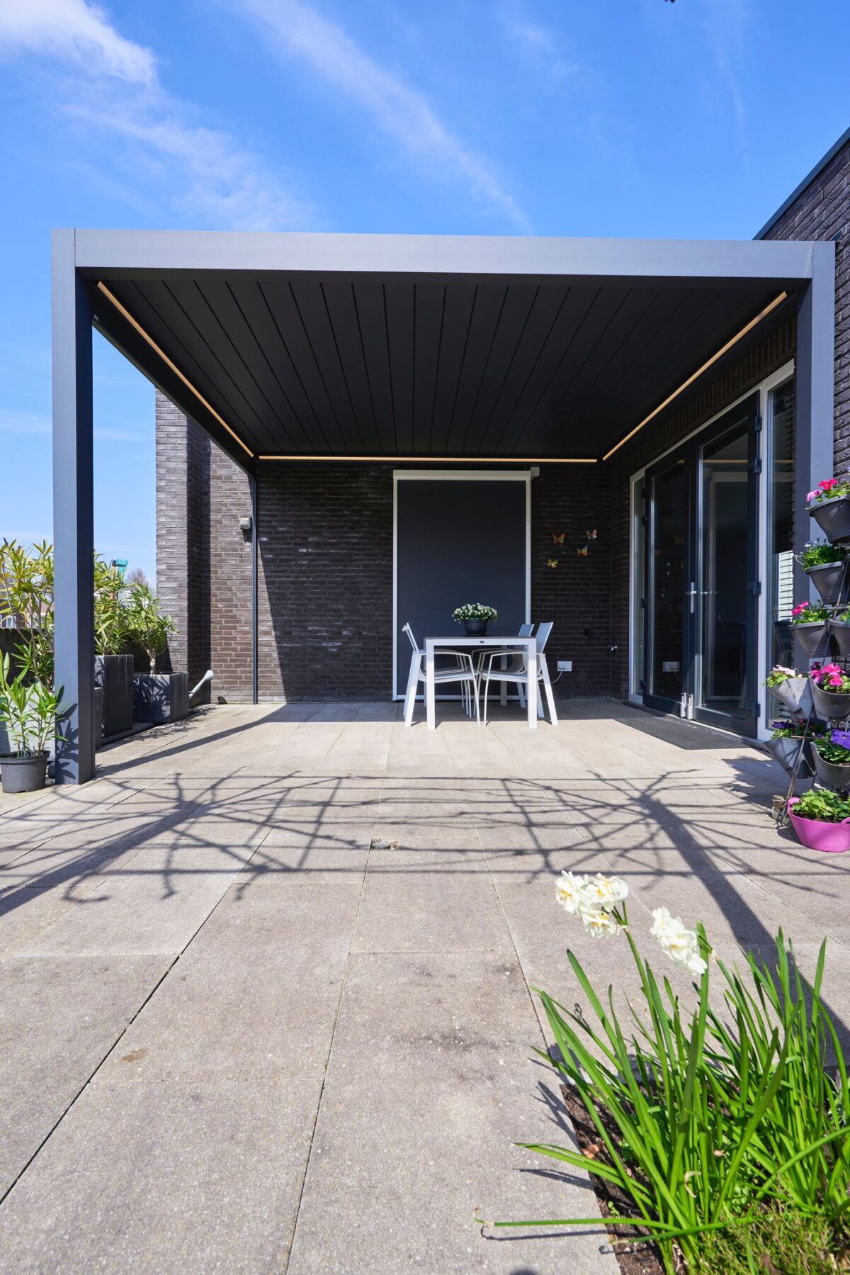 Een modern buitenterras met een strakke, donkere pergola die een eettafel en stoelen bedekt. Het gebied is omgeven door potplanten en bloemen, met een helderblauwe lucht erboven.