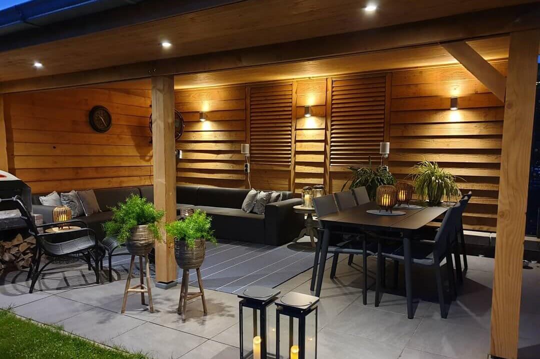 Gezellige patio met houten wanden en plafond, met een sectionele bank en eethoek. De ruimte wordt verlicht door warme sfeerverlichting. Planten en een grill dragen bij aan de uitnodigende sfeer.