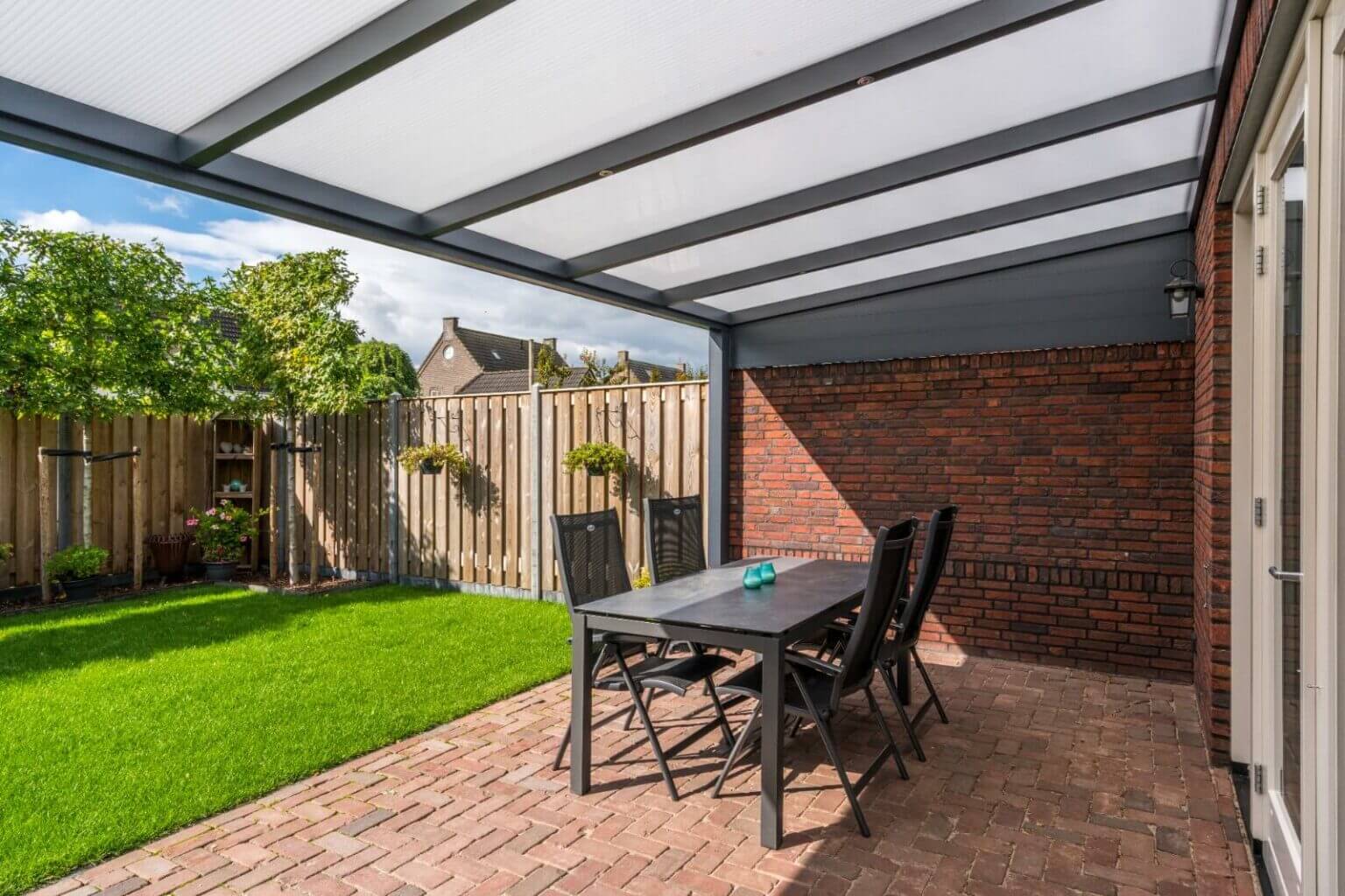 Overdekt terras met een donkere tafel en vier stoelen op een bakstenen vloer. Het gebied kijkt uit op een groen gazon met potplanten tegen een houten hek. Een bakstenen muur aan de rechterkant leidt naar een deur. De lucht is gedeeltelijk bewolkt.