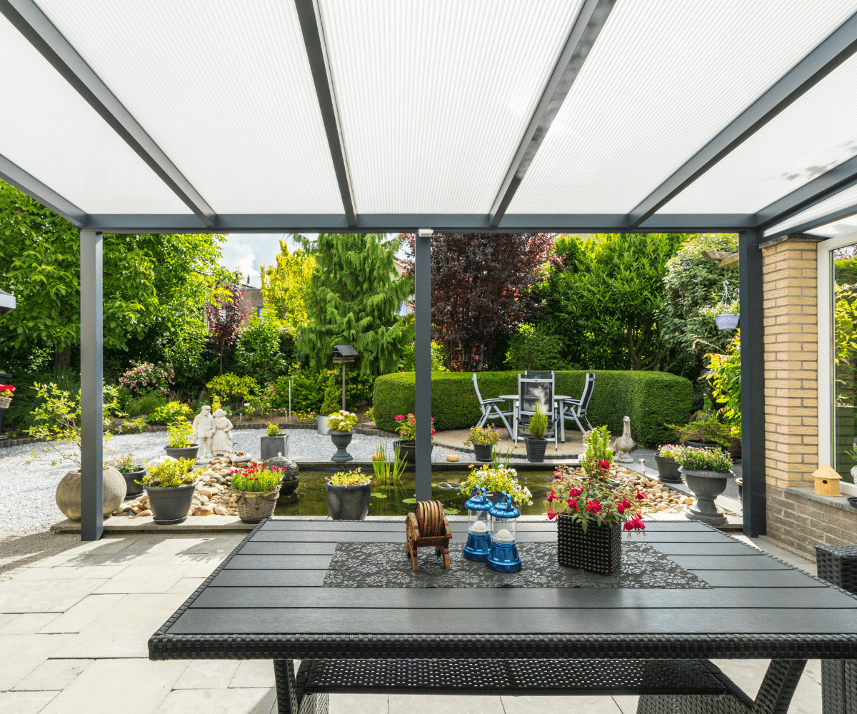 Overdekte patio met een zwarte tafel, decoratieve items en stoelen met uitzicht op een weelderige tuin. De tuin heeft potplanten, een kleine vijver en zitplaatsen buiten. De ruimte is omgeven door groen en heeft een serene, uitnodigende sfeer.