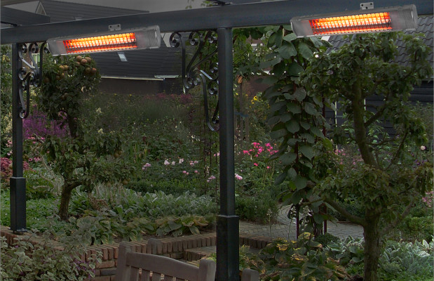 Buitenterras met hangende heaters aan een pergola, omgeven door weelderig groen en diverse bloeiende planten. Een houten bank is gedeeltelijk zichtbaar op de voorgrond.