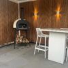 Buitenpatio met een houtgestookte pizzaoven naast een stapel brandhout tegen een houten lattenwand. Twee moderne witte barkrukken zijn aan een hoge witte tafel geplaatst. Warme wandlampen verlichten de ruimte en versterken de gezellige sfeer.