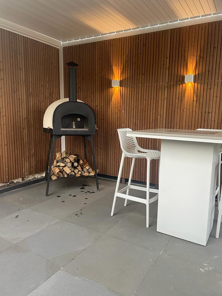 Buitenpatio met een houtgestookte pizzaoven naast een stapel brandhout tegen een houten lattenwand. Twee moderne witte barkrukken zijn aan een hoge witte tafel geplaatst. Warme wandlampen verlichten de ruimte en versterken de gezellige sfeer.