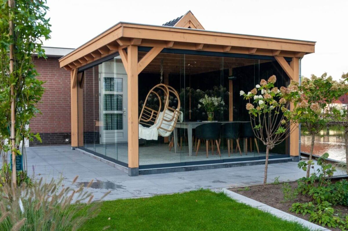 Een eigentijdse patio met de Houten overkapping Classic met glazen wanden. Binnen vindt u een hangstoel en een tafel met zwarte stoelen. De patio grenst aan een goed onderhouden tuin, compleet met gras, planten en een geplaveid wandelpad.