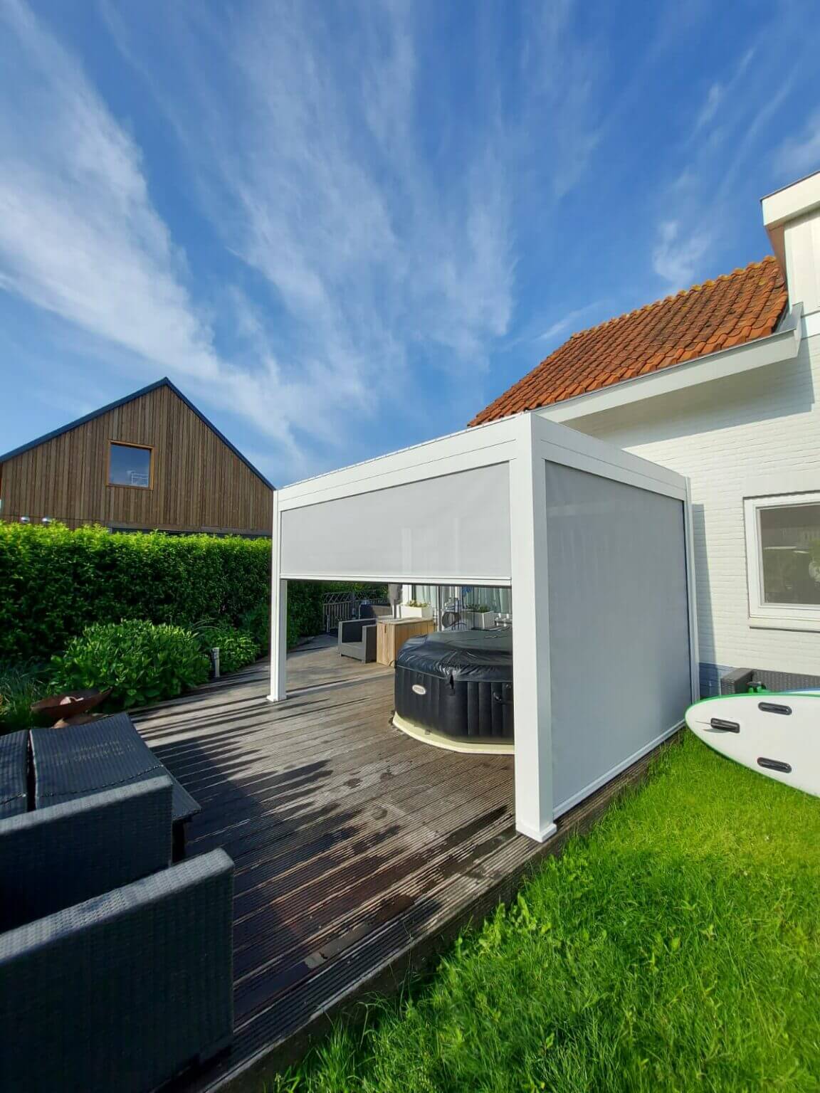 Een patio met een moderne witte pergola boven een ronde hottub, op een houten dek. Er staat zwart tuinmeubilair in de buurt. De lucht is helder met dunne wolken en er zijn groene gazons en een houten huis op de achtergrond.