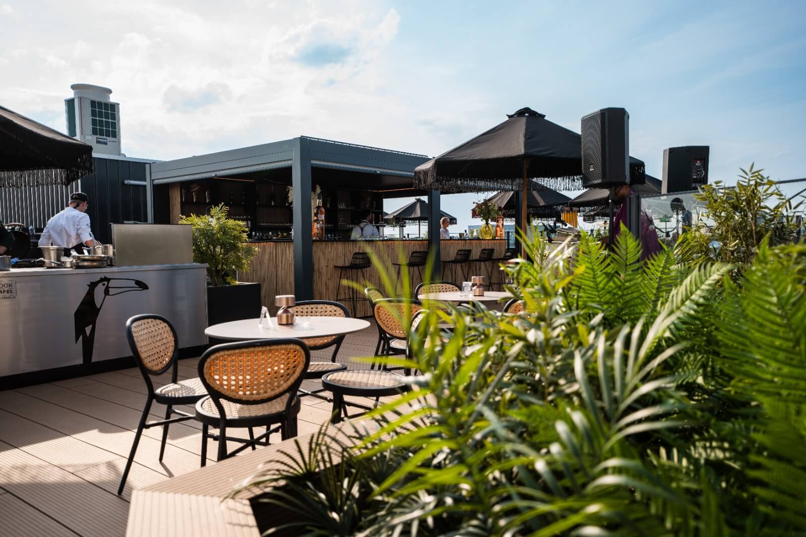 Dakterras met tafels en stoelen omgeven door groen. Een bar en een chef-kok die aan het werk is bij een kookstation zijn zichtbaar. Parasols bieden schaduw en de lucht is helder met verspreide wolken.