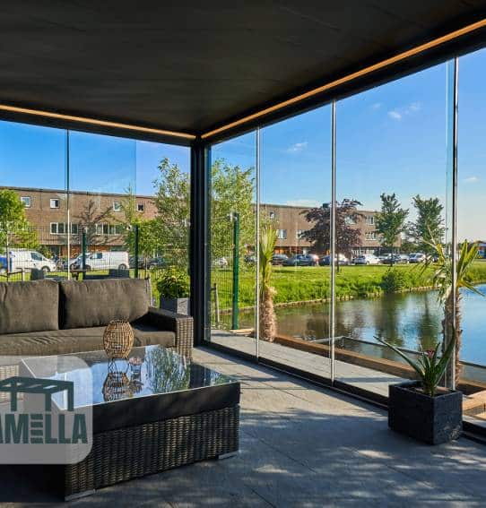 Een moderne serre met glazen wanden en een nachhaltige aluminiumüberdachung biedt uitzicht op de buitenlucht, met bomen en een vijver. De kamer heeft een donkere bank, een salontafel met glazen blad en potplanten, wat een gezellige en eigentijdse sfeer creëert.