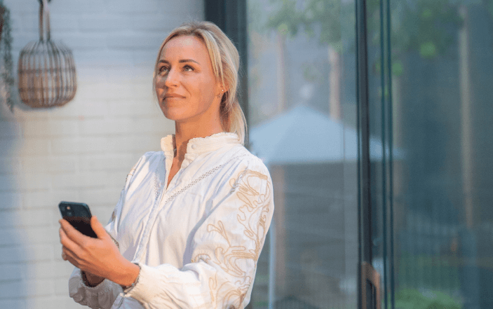 Een persoon met licht haar, gekleed in een wit shirt, staat binnen met een smartphone in zijn hand. Ze kijken omhoog met een lichte glimlach. Een raam aan hun rechterkant laat een wazig zicht op de buitenwereld zien.