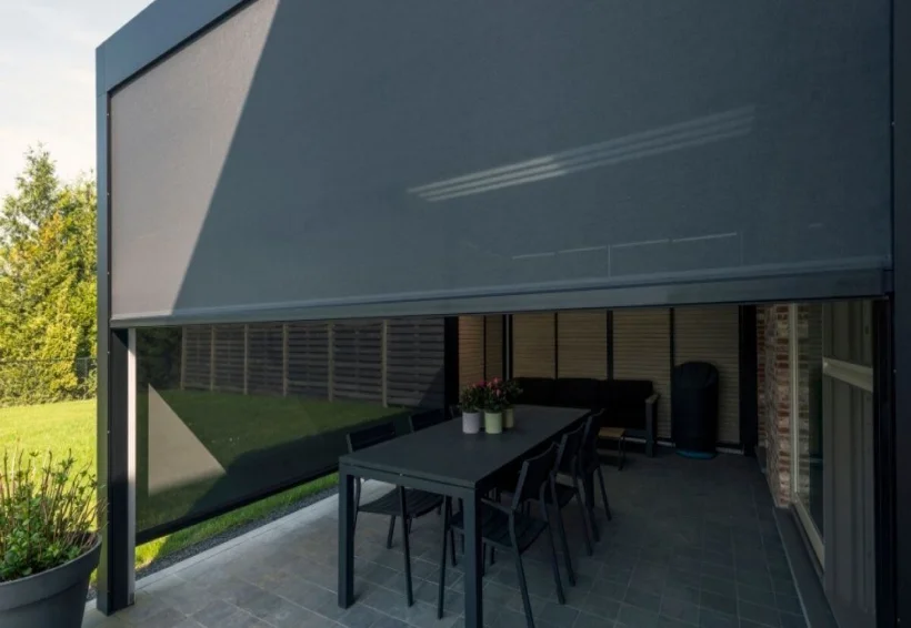 Een modern terras met een gedeeltelijk neergelaten grijs uitschuifbaar scherm dat een eettafel met zes stoelen beschaduwt. Potplanten staan op de tafel en in de hoek; buiten is groen gras zichtbaar.