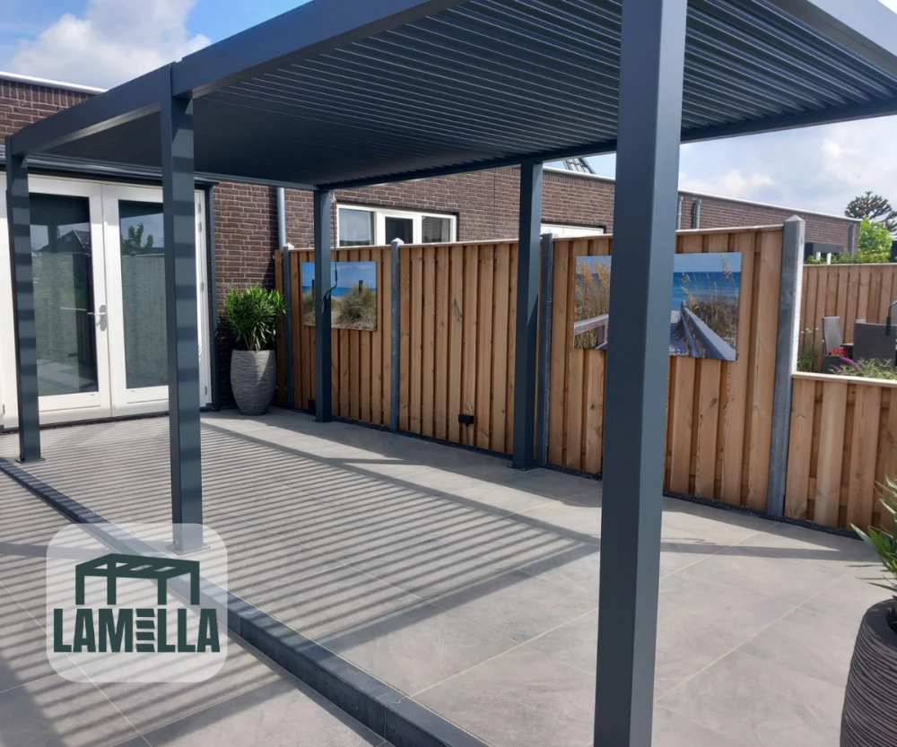 Moderne patio met een grijze pergola die gestreepte schaduwen werpt op een betegelde vloer. Houten hek met ingelijste landschapsfotos. Potplanten bij glazen deuren. Lamella-logo in de linkerbenedenhoek.