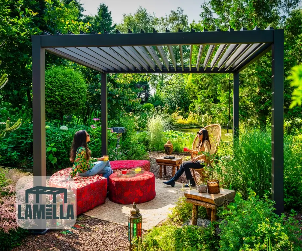 Twee mensen zitten onder een moderne pergola in een weelderige tuin, omringd door rode, gepatroneerde ronde zitplaatsen en houten tafels. Een lantaarn rust op de grond. Zonlicht filtert door bomen en werpt schaduwen. Een Lamella-logo staat in de linkerbenedenhoek.