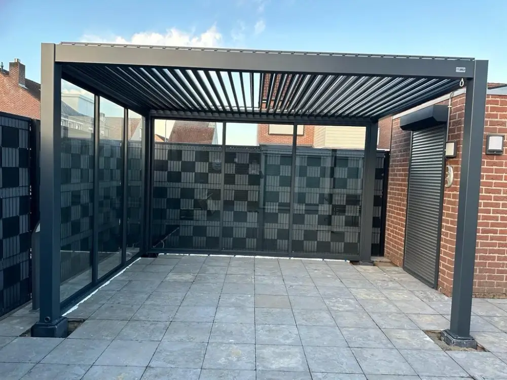 Een moderne buitenpergola met een lamellendak staat op een betegeld terras. Glazen wanden omsluiten de ruimte gedeeltelijk, waardoor er zicht is op de omliggende bakstenen gebouwen en een geblokte privacyomheining. De lucht erboven is helder en blauw.