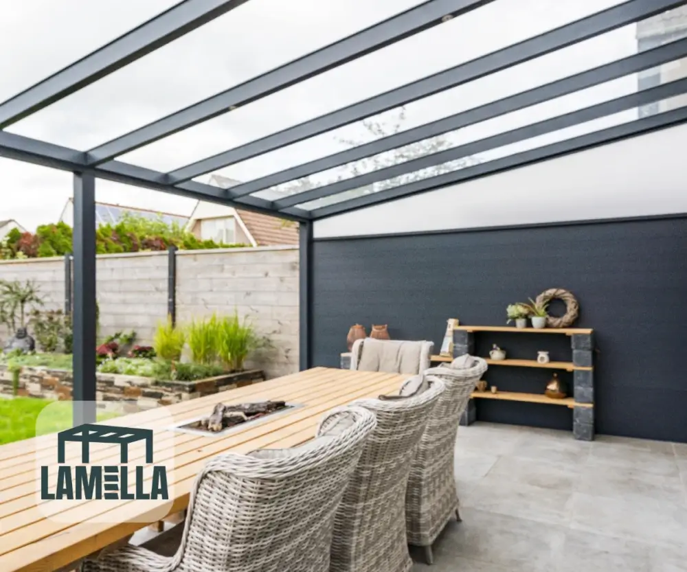 Buitenpatio met een pergola met glazen dak en een lange houten eettafel omringd door rieten stoelen. Links is een tuin zichtbaar en rechts is een stenen plank met decoratieve items. Lamella-logo in de linkerbenedenhoek.