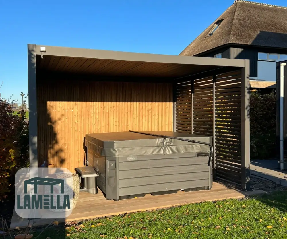 Buitenjacuzzi onder een moderne pergola met houten lattenwanden, geplaatst op een houten dek. De constructie staat in een achtertuin met gedeeltelijk zonlicht. Op de achtergrond is een huis met een rieten dak zichtbaar.