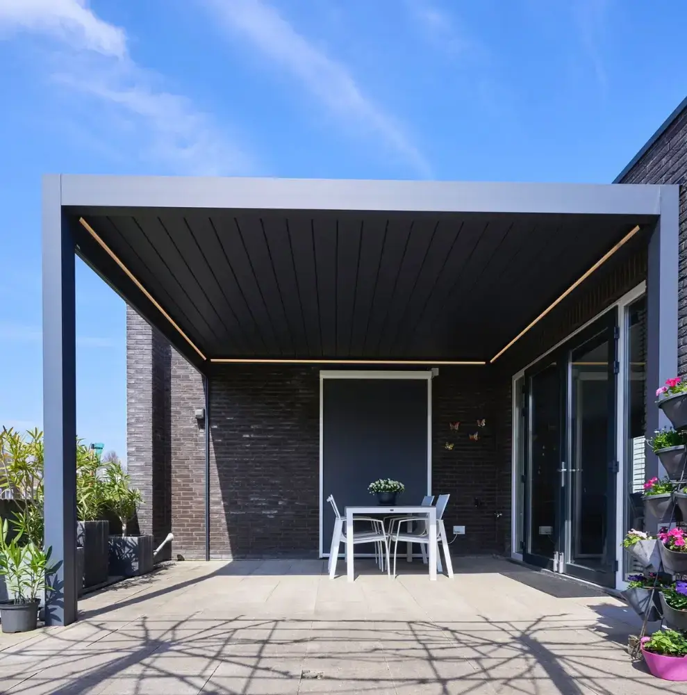 Een moderne patio met een zwarte pergola en een kleine witte tafel met stoelen eronder. Het gebied is omringd door potplanten en de achtergrond heeft een glazen schuifdeur. De lucht is helder en blauw.