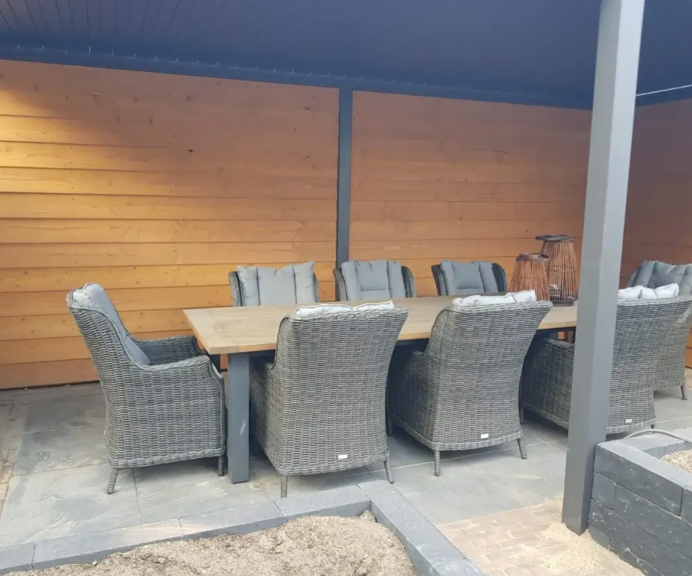 Buitenpatio met een grote houten eettafel omringd door acht gevlochten rotanstoelen met kussens. Het gebied heeft een houten muur, een betegelde vloer en lantaarns op de tafel, wat een gezellige en uitnodigende sfeer creëert.