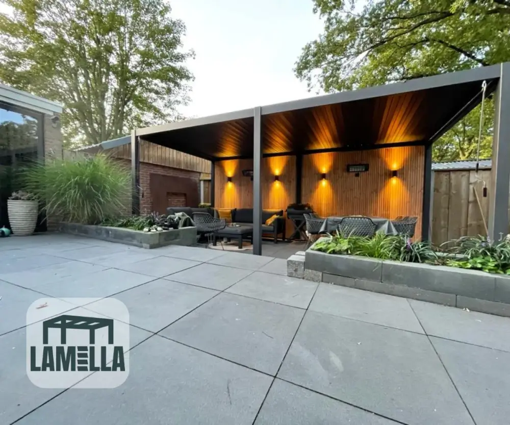 Een modern buitenterras met een houten pergola die een zitgedeelte bedekt. Warme lichten verlichten de met hout beklede muur en er is weelderig groen rondom. Het logo Lamella is zichtbaar in de linkerbenedenhoek.