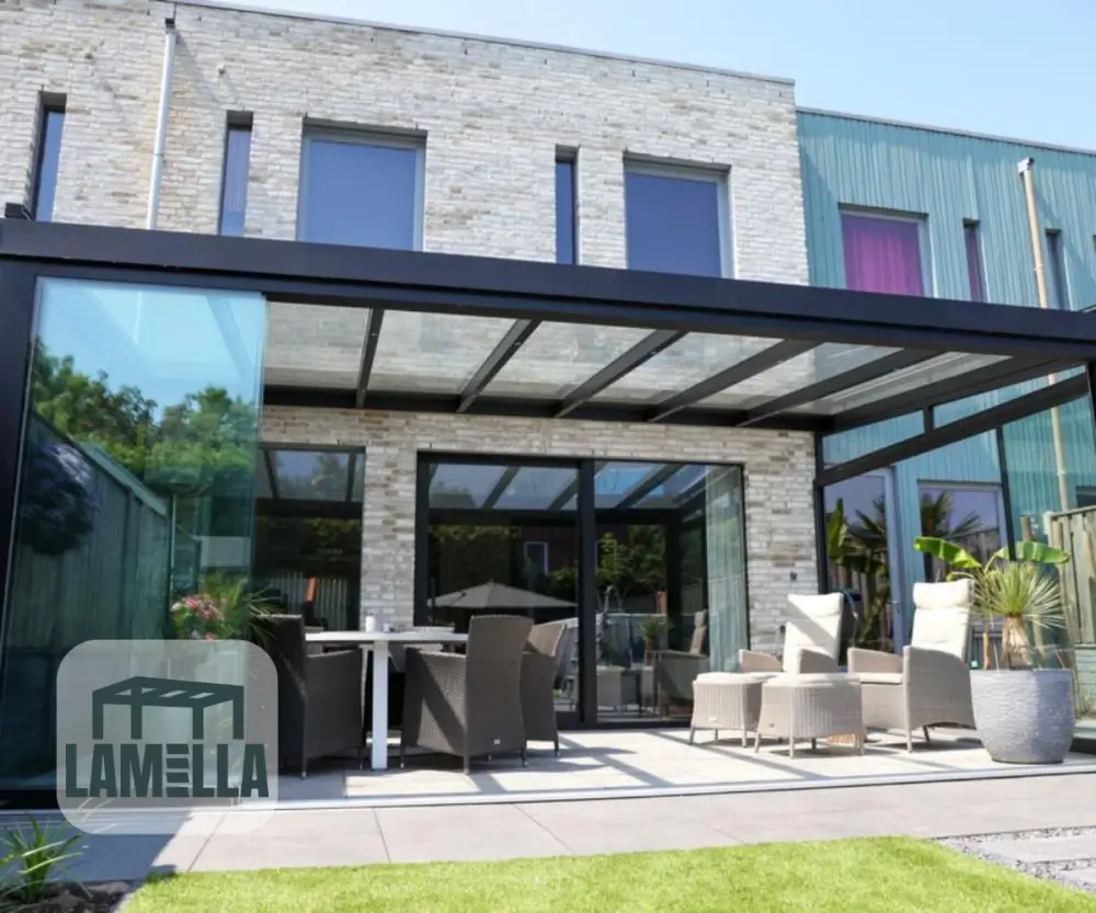 Modern huis met een glazen luifel die een patio bedekt. De patio is voorzien van rieten meubels, waaronder stoelen en een tafel. De gevel van het huis combineert bakstenen en blauwgroene panelen. Het weer is zonnig en de tuin is groen met een logo in de linkerbenedenhoek.