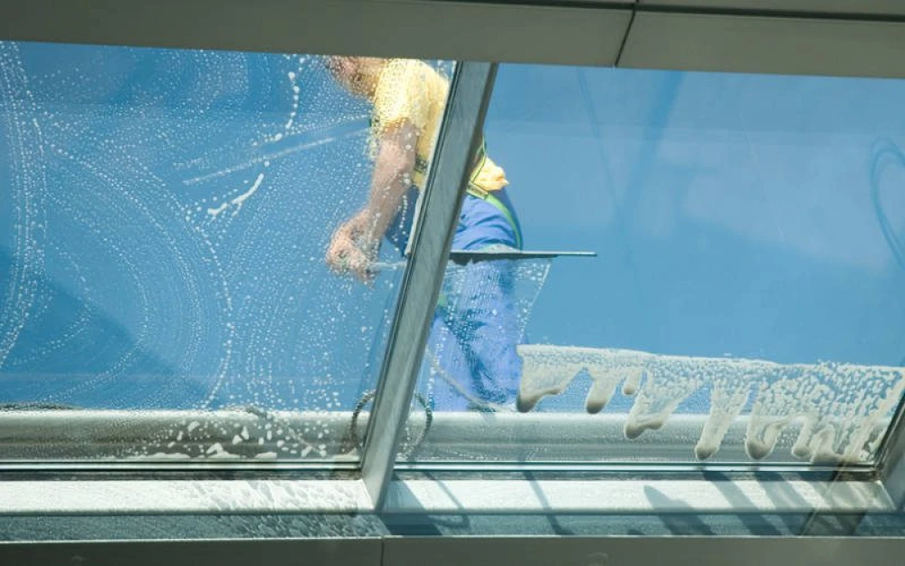 Een glazenwasser in geel en blauw gewaad reinigt een groot glazen raam van buitenaf. Het raam is gedeeltelijk bedekt met zeepsop, met zichtbare waterstrepen. De helderblauwe lucht is op de achtergrond te zien.