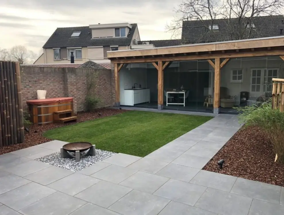 Moderne achtertuin met een vierkant stuk gras, omgeven door grijze tegels. Kenmerken zijn onder andere een vuurplaats op kiezelstenen, een houten hut met glazen wanden en een hot tub. Bakstenen muur en aangrenzende huizen zijn zichtbaar op de achtergrond.