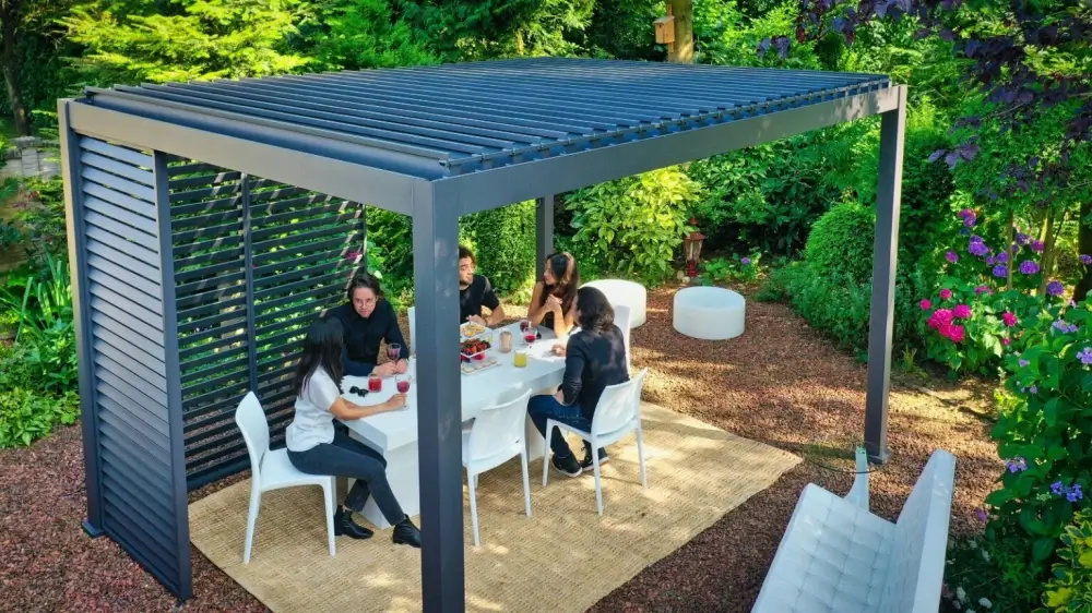 Mensen zitten rond een witte tafel onder een moderne metalen pergola in een weelderige tuin. De pergola heeft een lamellendak en zijkanten. De scène omvat witte stoelen, drankjes, snacks en groen met kleurrijke hortensias. Een geweven kleed ligt onder de tafel.