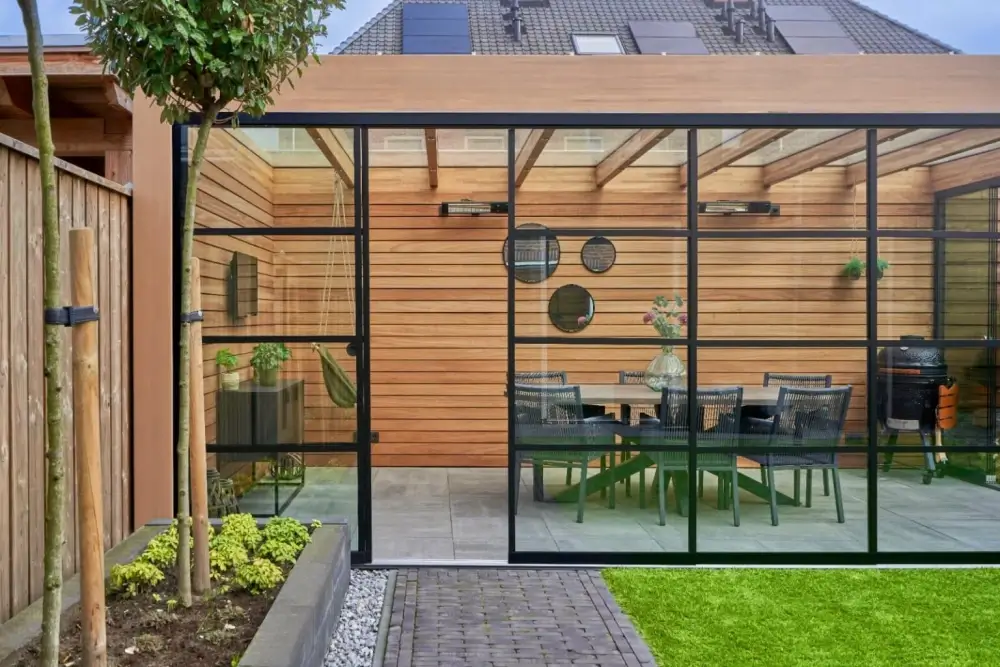 Moderne glazen patio met houten lattenwanden en plafond. Binnen is er een eettafel met stoelen, decoratieve ronde spiegels en een vaas met bloemen. Buiten is er een groen gazon en een geplaveid pad.