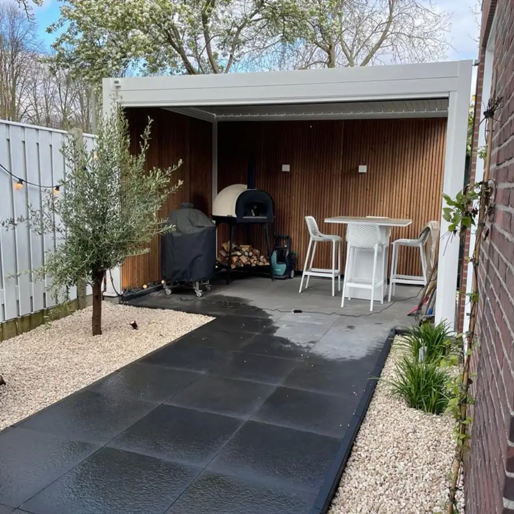 Een modern buitenterras heeft een overdekte zithoek met een hoge tafel en krukken, een pizzaoven, een grill en brandhout. De ruimte wordt begrensd door een bakstenen muur en een wit hek, met een boom en kleine planten op een grindbed.