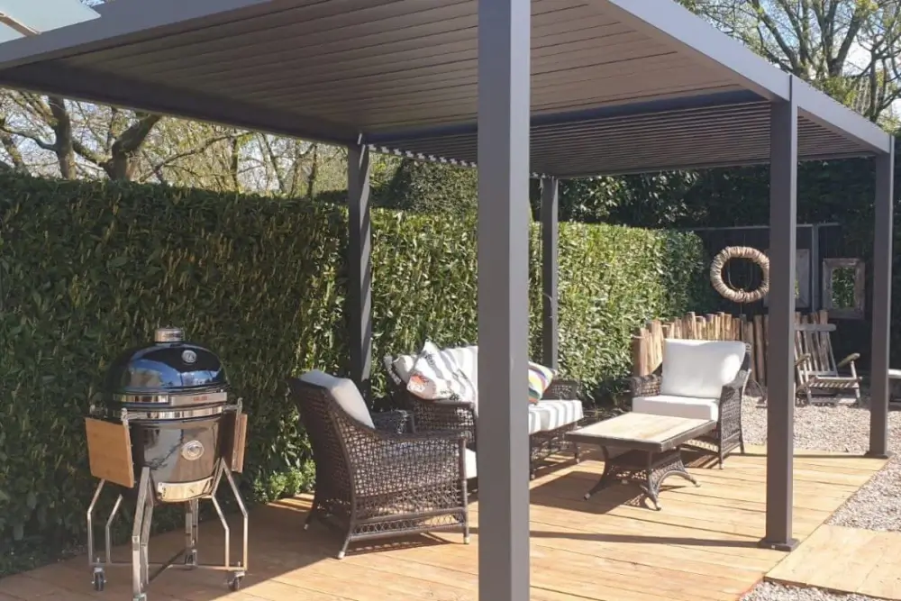 Buitenpatio met een moderne pergola boven een terras. Er is een ronde grill aan de linkerkant en een zithoek met een rieten bank en stoelen, gestreepte kussens en een salontafel. Een hoge heg omringt de ruimte en er hangt een krans aan de rechtermuur.