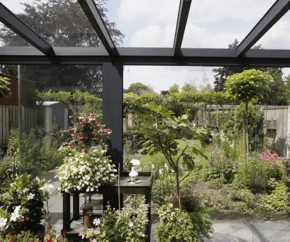 Een serre met glazen wanden en plafond kijkt uit op een weelderige tuin. Binnen staan er potplanten en een kleine tafel. Buiten heeft de tuin verschillende planten en bomen onder een bewolkte lucht. Er loopt een hek langs de rand van de tuin.