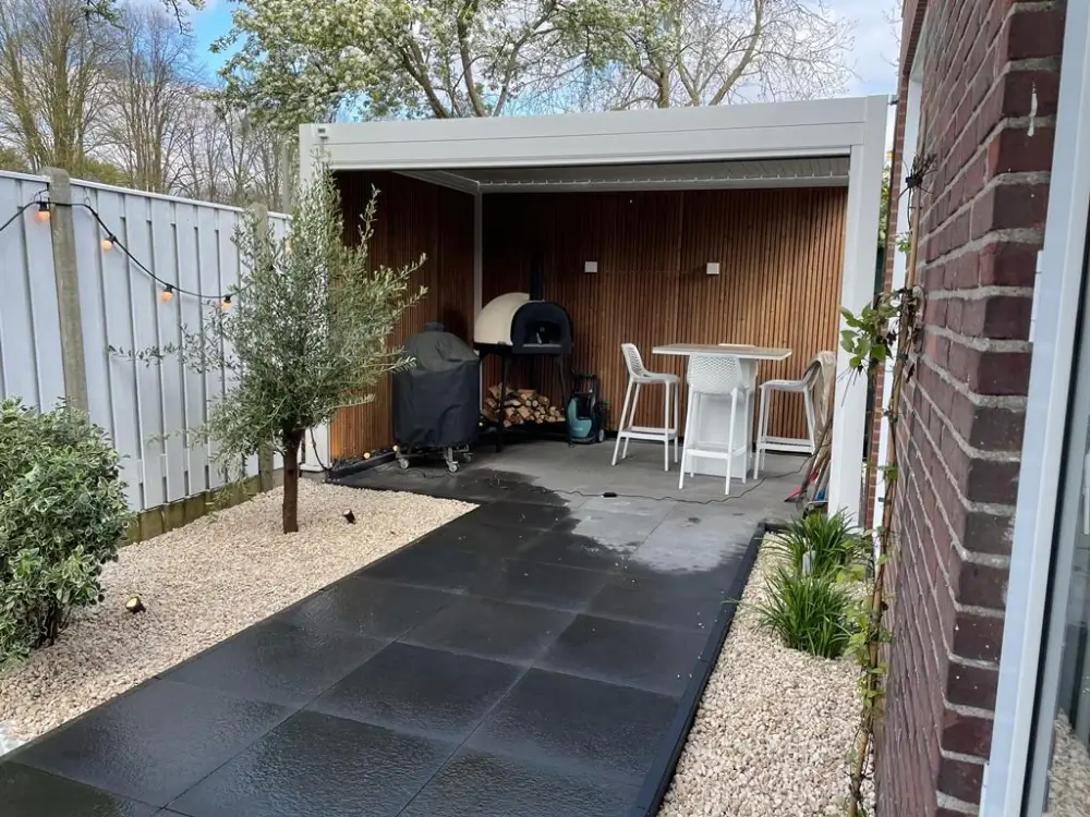 Een modern buitenterras met een houten pergola met een pizzaoven, stoelen en een tafel. Het gebied is omgeven door een wit hek en groen, met grote zwarte tegels op de grond en een olijfboom in de buurt.