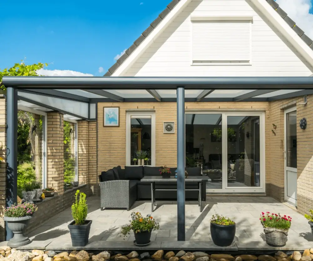 Een bakstenen huis heeft een moderne patio met een zwarte metalen pergola die een zitgedeelte bedekt. Er staan potplanten rondom de ruimte, glazen deuren die naar binnen leiden en een helderblauwe lucht erboven.