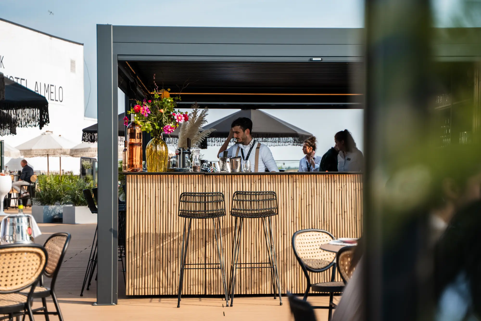 Een man staat bij een buitenbar met een bamboe front, prachtig versierd met kleurrijke bloemen. Twee barkrukken en terrastafels nodigen je uit om te zitten en te genieten van de buitenkeuken-vibe. De heldere, zonnige ambiance wordt aangevuld met parasols en weelderige planten op de achtergrond.