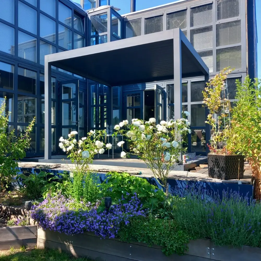 Moderne, kubusvormige metalen prieel in een tuin met verschillende planten en witte bloemen. Daarachter is een gebouw met meerdere verdiepingen en grote glazen ramen zichtbaar. Op de voorgrond weelderig groen en paarse bloemen onder een zonnige, helderblauwe lucht.